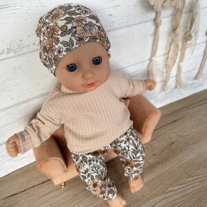 May include: A doll wearing a floral print hat, a peach-colored sweater, and floral print pants. The doll is sitting on a small brown chair.