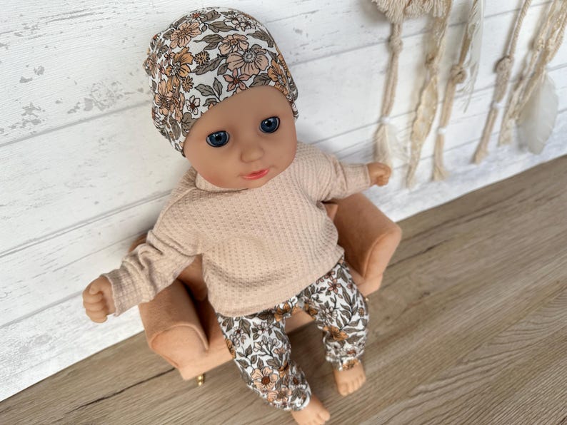 May include: A doll wearing a floral print hat, a peach-colored sweater, and floral print pants. The doll is sitting on a small brown chair.