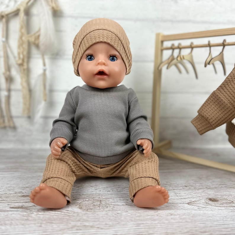 May include: A doll with blue eyes wearing a tan waffle-knit beanie, a grey long-sleeved ribbed shirt, and matching tan waffle-knit trousers. The doll has a neutral expression and is seated in a forward-facing position with bare feet visible.