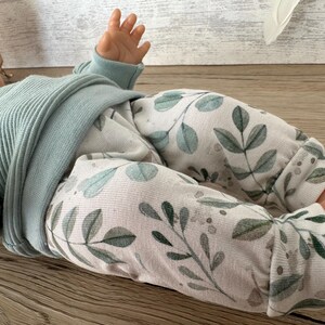 May include: A long-sleeved ribbed shirt in a solid light blue colour paired with white trousers featuring a repeating pattern of green and grey leaves and small dots. The garment set is shown on a doll, highlighting the textured fabric and botanical print.