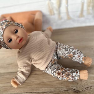 May include: A doll wearing a beige long-sleeved shirt and floral print pants. The doll is wearing a floral print headband.