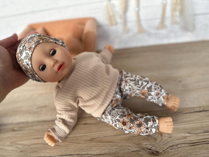 May include: A doll wearing a beige long-sleeved shirt and floral print pants. The doll is wearing a floral print headband.