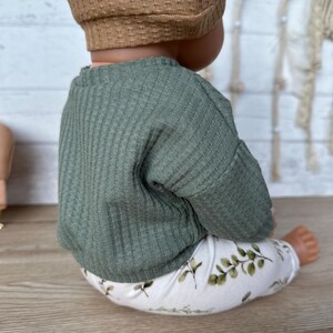 May include: A green ribbed sweater and white leggings with a leafy print. The outfit is worn by a doll.