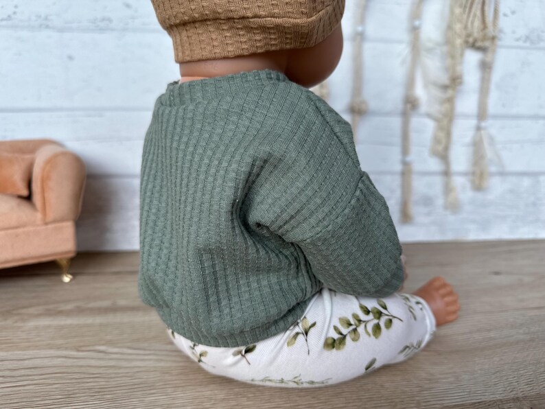 May include: A green ribbed sweater and white leggings with a leafy print. The outfit is worn by a doll.