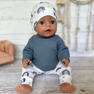 May include: A doll wearing a white beanie with a blue rainbow pattern, a solid dark blue long-sleeved waffle-knit shirt, and white pants featuring a matching blue rainbow pattern. The doll has blue eyes and a neutral expression, sitting upright.