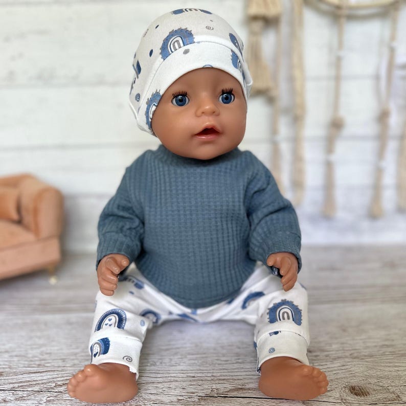 May include: A doll wearing a white beanie with a blue rainbow pattern, a solid dark blue long-sleeved waffle-knit shirt, and white pants featuring a matching blue rainbow pattern. The doll has blue eyes and a neutral expression, sitting upright.