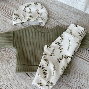 K&ouml;nnte beinhalten: Ein gr&uuml;ner Waffelstrickpullover, eine wei&szlig;e Hose mit gr&uuml;nem Blattmuster und eine wei&szlig;e M&uuml;tze mit gr&uuml;nem Blattmuster.