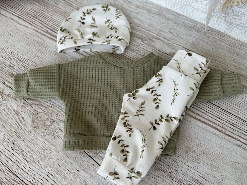 K&ouml;nnte beinhalten: Ein gr&uuml;ner Waffelstrickpullover, eine wei&szlig;e Hose mit gr&uuml;nem Blattmuster und eine wei&szlig;e M&uuml;tze mit gr&uuml;nem Blattmuster.