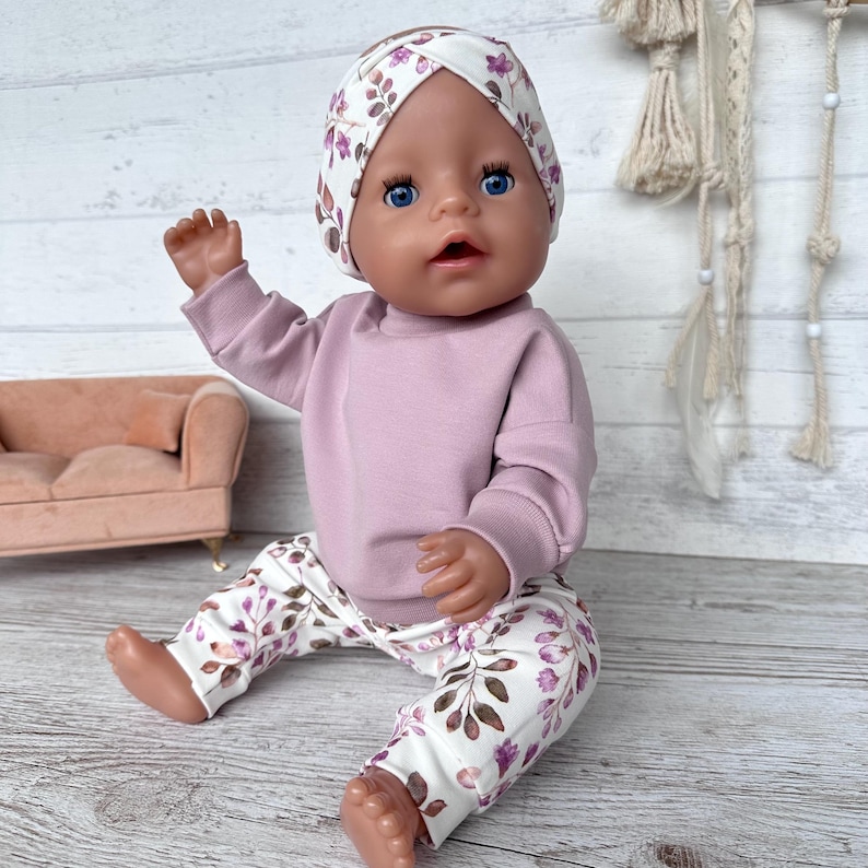 May include: A doll wearing a mauve long-sleeved sweatshirt, white leggings with a purple and brown leaf pattern, and a matching white headband with a knotted front detail. The doll has blue eyes and is positioned in a seated posture.
