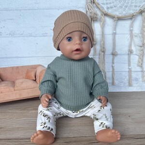 May include: A doll wearing a green waffle knit sweater, white pants with a green leaf pattern, and a brown beanie. The doll is sitting on a light brown couch.