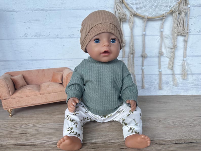 May include: A doll wearing a green waffle knit sweater, white pants with a green leaf pattern, and a brown beanie. The doll is sitting on a light brown couch.