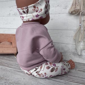 May include: A doll wearing a solid mauve long-sleeved sweatshirt paired with white leggings and a matching headband, both featuring a botanical print of purple flowers and brown leaves. The outfit is designed for a small doll figure.