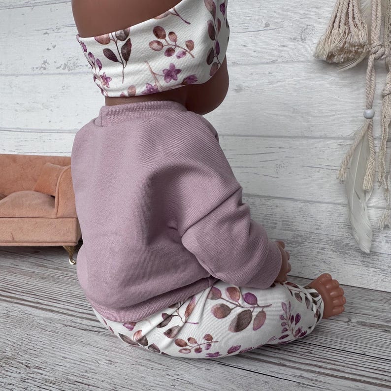 May include: A doll wearing a solid mauve long-sleeved sweatshirt paired with white leggings and a matching headband, both featuring a botanical print of purple flowers and brown leaves. The outfit is designed for a small doll figure.