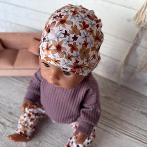 May include: A doll wearing a floral print beanie, a mauve waffle-knit long-sleeve shirt, and matching floral print trousers. The beanie features a white background with brown and orange flowers.