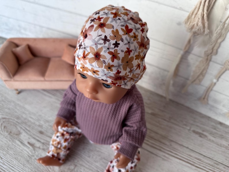 May include: A doll wearing a floral print beanie, a mauve waffle-knit long-sleeve shirt, and matching floral print trousers. The beanie features a white background with brown and orange flowers.