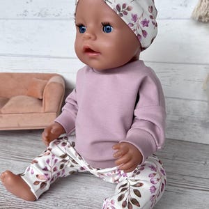 May include: A doll with blue eyes wearing a mauve long-sleeve sweatshirt, white leggings with a purple and brown floral leaf pattern, and a matching white headband with a twisted front featuring the same purple and brown floral leaf print.