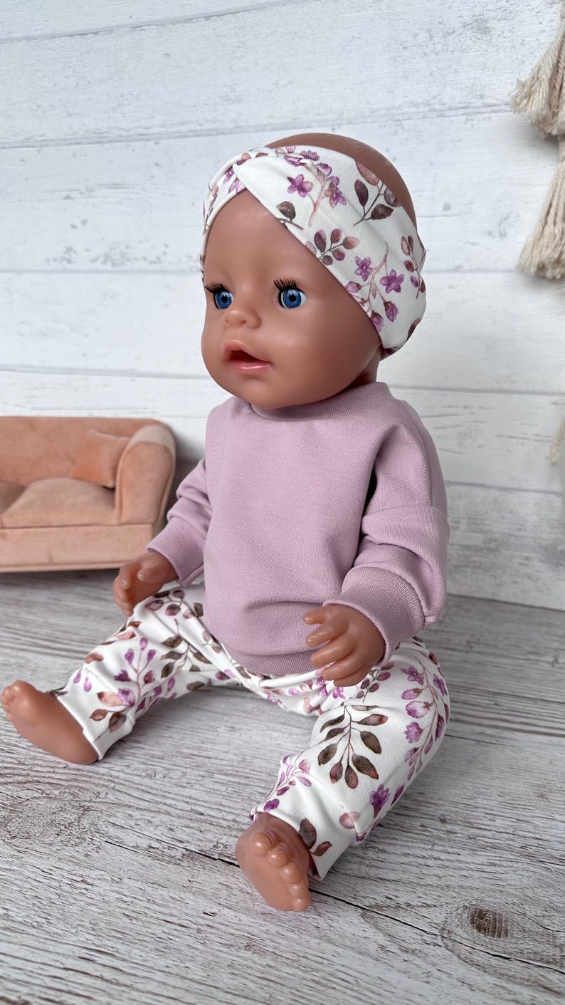 May include: A doll with blue eyes wearing a mauve long-sleeve sweatshirt, white leggings with a purple and brown floral leaf pattern, and a matching white headband with a twisted front featuring the same purple and brown floral leaf print.