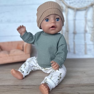 May include: A doll wearing a green waffle knit long-sleeve shirt, white pants with a green leaf pattern, and a brown knit beanie.
