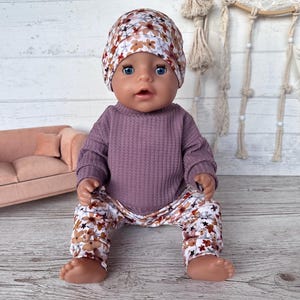 May include: A doll dressed in a mauve waffle-knit top, floral print trousers, and a matching beanie. The trousers and beanie have a pattern of brown and white flowers. The doll is sitting on a wooden surface.