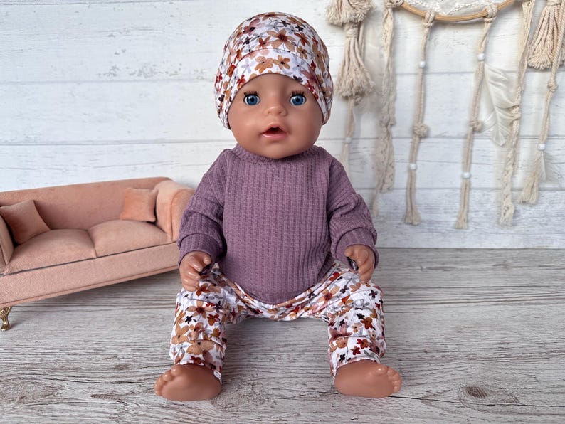May include: A doll dressed in a mauve waffle-knit top, floral print trousers, and a matching beanie. The trousers and beanie have a pattern of brown and white flowers. The doll is sitting on a wooden surface.