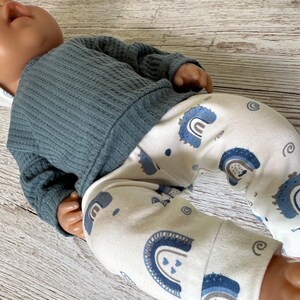 May include: A doll wearing a long-sleeved, dark blue, textured waffle-knit shirt and white leggings featuring a repeating pattern of blue rainbows and small swirls. The doll has a smooth, light-toned complexion and is positioned at an angle.