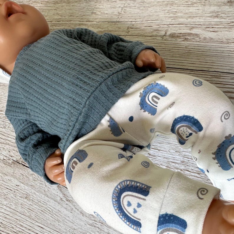 May include: A doll wearing a long-sleeved, dark blue, textured waffle-knit shirt and white leggings featuring a repeating pattern of blue rainbows and small swirls. The doll has a smooth, light-toned complexion and is positioned at an angle.