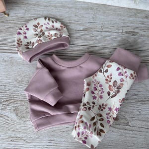 May include: A three-piece clothing set consisting of a solid dusty rose long-sleeve sweatshirt, matching solid dusty rose pants, and a beanie. The beanie and one leg of the pants feature a white fabric with a botanical print of brown and purple leaves.
