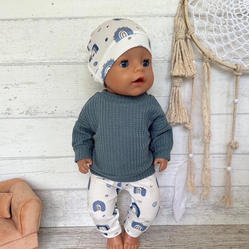May include: A doll wearing a long-sleeve dark blue waffle-knit shirt, white pants with a blue rainbow pattern, and a matching white beanie with a blue rainbow print. The doll has blue eyes and a neutral expression, standing upright in a set.