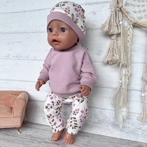 May include: A doll wearing a long-sleeved mauve sweatshirt, matching mauve and white floral print leggings, and a coordinating mauve and white floral print beanie hat. The doll has blue eyes and a neutral expression, dressed in a three-piece outfit.