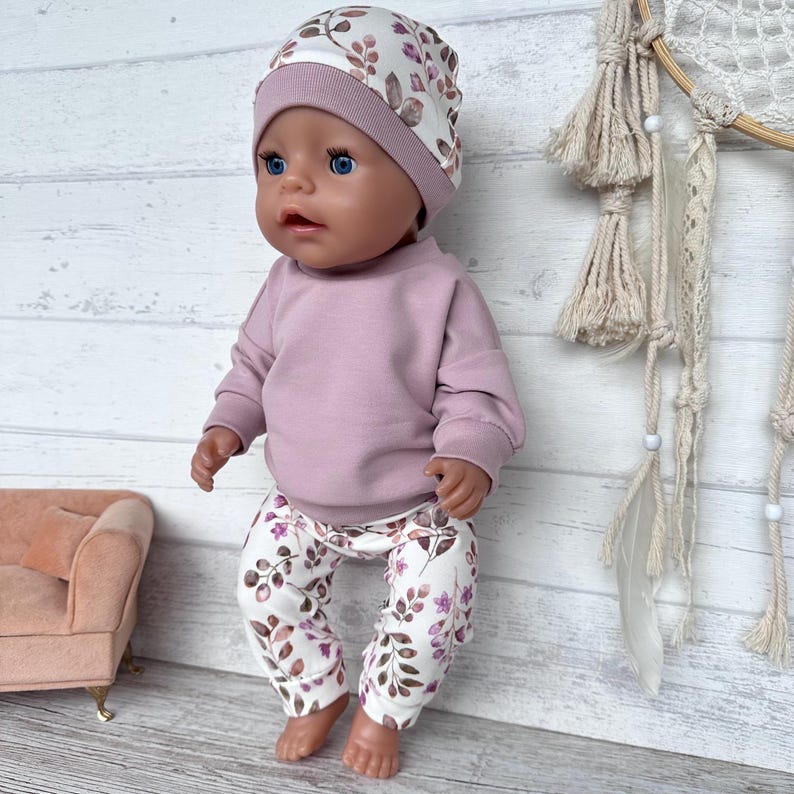 May include: A doll wearing a long-sleeved mauve sweatshirt, matching mauve and white floral print leggings, and a coordinating mauve and white floral print beanie hat. The doll has blue eyes and a neutral expression, dressed in a three-piece outfit.