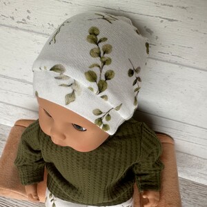 May include: A white baby hat with a green leafy pattern. The hat is made of soft, stretchy fabric and is perfect for a newborn or infant.