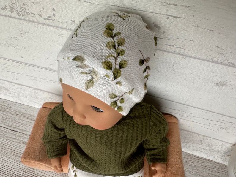 May include: A white baby hat with a green leafy pattern. The hat is made of soft, stretchy fabric and is perfect for a newborn or infant.
