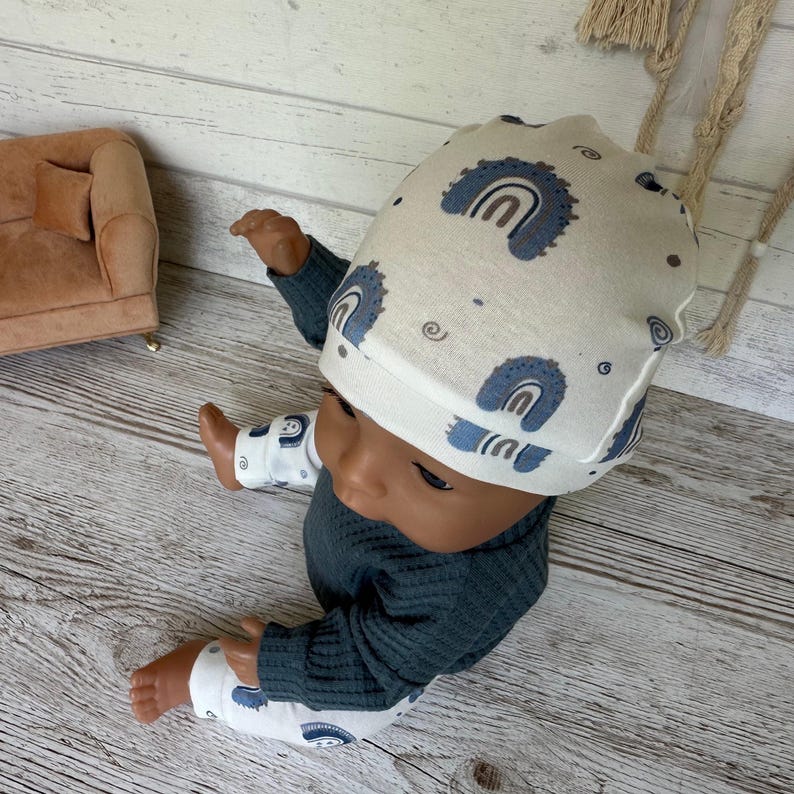 May include: A doll wearing a cream-colored beanie with a blue rainbow pattern, a dark gray long-sleeved ribbed shirt, and white leggings featuring a matching blue rainbow print. The doll is positioned in a seated posture on a light wood surface.