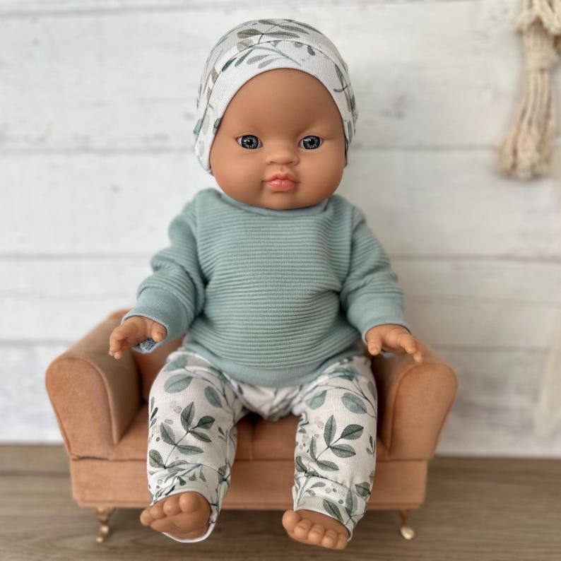 May include: A baby doll with a dark skin tone, wearing a white beanie with a green leaf pattern, a sage green ribbed long-sleeve shirt, and matching white trousers with a green leaf print. The doll is seated on a small, tan-coloured armchair.