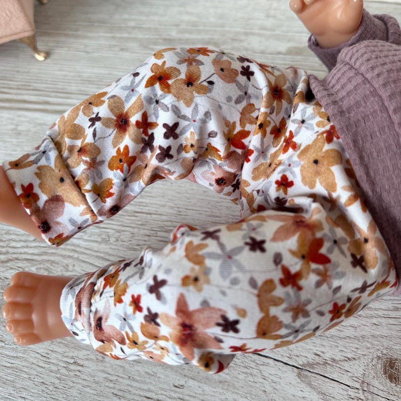 May include: Baby trousers featuring a floral design in brown, orange, and grey on a white base. The trousers are part of a baby outfit, with a purple long-sleeved top visible.