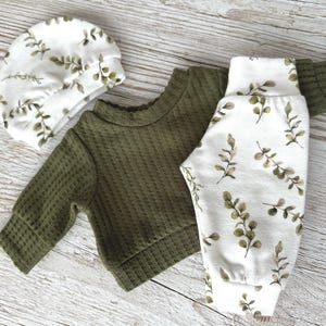 May include: A baby outfit set featuring a green waffle knit long-sleeved top and white trousers with a green leaf pattern. The set also includes a white hat with a green leaf pattern.
