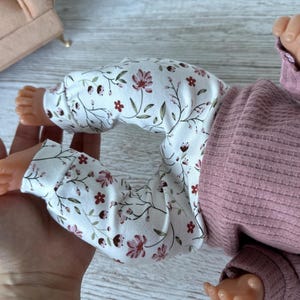 May include: A pair of white leggings featuring a delicate floral pattern with small pink and dark red flowers and green stems, paired with a solid mauve-colored long-sleeved ribbed shirt. The clothing is worn by a small doll with visible feet.