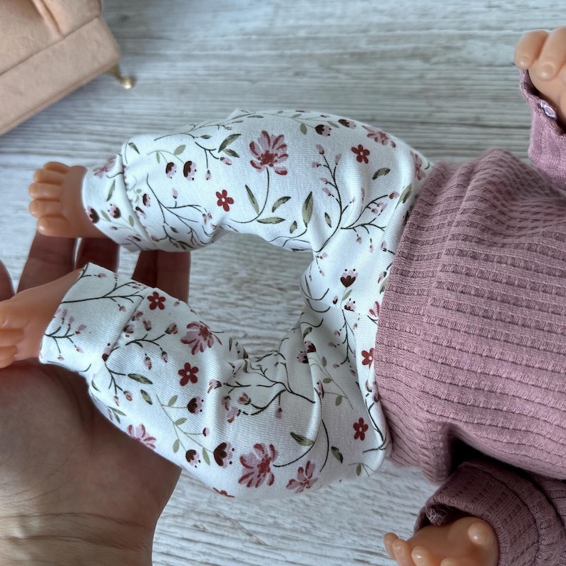 May include: A pair of white leggings featuring a delicate floral pattern with small pink and dark red flowers and green stems, paired with a solid mauve-colored long-sleeved ribbed shirt. The clothing is worn by a small doll with visible feet.