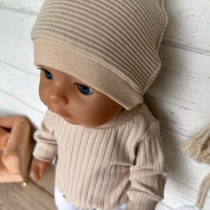 Puede incluir: Un conjunto de gorro y camisa de manga larga de punto de canal&eacute; beige para una mu&ntilde;eca. La mu&ntilde;eca tiene ojos azules y una tez marr&oacute;n claro.