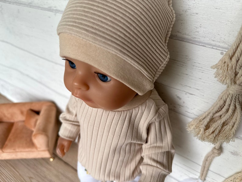 Puede incluir: Un conjunto de gorro y camisa de manga larga de punto de canal&eacute; beige para una mu&ntilde;eca. La mu&ntilde;eca tiene ojos azules y una tez marr&oacute;n claro.