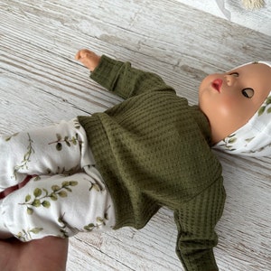 May include: A doll wearing a green waffle knit long-sleeved shirt, white trousers with a leafy print, and a white hat with a leafy print.