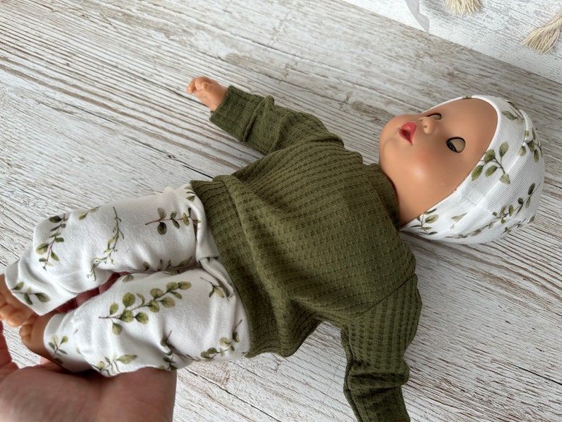 May include: A doll wearing a green waffle knit long-sleeved shirt, white trousers with a leafy print, and a white hat with a leafy print.