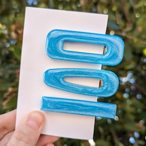 Puede incluir: Tres pinzas para el pelo azules con un acabado brillante. Las pinzas son de diferentes formas y tamaños. La pinza más grande es un rectángulo con un borde redondeado. La pinza del medio es un rectángulo redondeado. La pinza más pequeña es un rectángulo delgado.