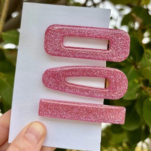 Puede incluir: Tres pinzas para el pelo rosas con purpurina en forma rectangular, ovalada y de barra se muestran en una tarjeta blanca. Las pinzas tienen un acabado brillante y cierres metálicos dorados. El fondo es follaje verde borroso.