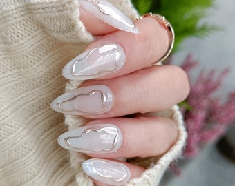 May include: Close-up of a hand with long, almond-shaped nails. The nails are painted a light, milky white with a silver outline design. The design resembles abstract shapes or clouds.