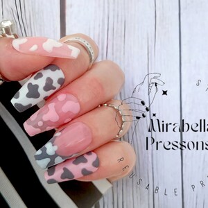 Könnte beinhalten: Press-on-Nägel mit Kuhmuster und rosafarbenem Muster. Die Nägel sind lang und haben einen glänzenden Finish. Die Nägel werden auf einer Hand mit weißem Hintergrund gezeigt.