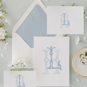 Könnte beinhalten: Briefpapier-Set mit einem weißen Umschlag mit hellblauem Innenfutter und passenden Karten. Jede Karte zeigt ein hellblaues Monogramm-Design. Das Set ist mit weißen Blumen und einer kleinen Schale mit einer Blumenmarke arrangiert.