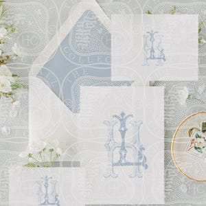 May include: A stationery set featuring white paper cards and an envelope with a blue patterned liner. Each card displays a blue decorative monogram. The set is arranged with white floral accents and a small dish containing a postage stamp.