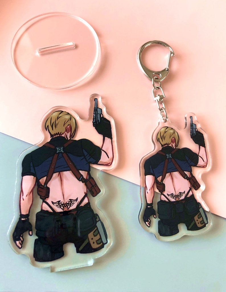 Op de afbeelding: Een transparante acryl sleutelhanger met een cartoonfiguur dat twee pistolen vasthoudt. Het personage draagt een zwart shirt, een zwarte broek en een bruine riem. Het personage heeft blond haar en een tatoeage op zijn rug.