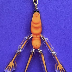 May include: A clear acrylic keychain featuring an orange cartoon character with a segmented body, sunglasses, and three pronged appendages. The keychain has a gold-colored chain and clasp, set against a purple background.